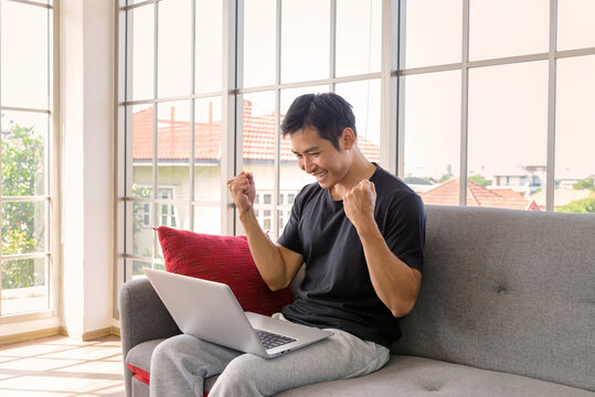 Happy Asian Man Sitting On Cozy Couch. His Punching The Air While Working With Laptop Computer. Successful Concept