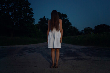 an adult girl in a white dress with long hair turned over her face stands at a road intersection in...