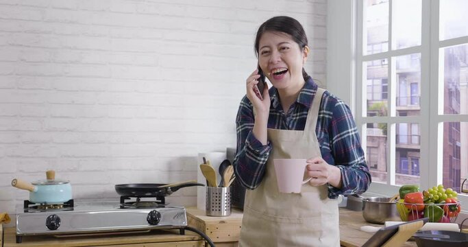Young relax korean woman wearing apron talking on cellphone in modern wooden style kitchen. beautiful lady laughing holding cup of morning coffee and standing in cooking place in cozy apartment.