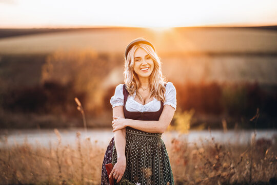 Pretty blonde girl in dirndl, standing outdoors in the field, holding bouquet of a field flowers.