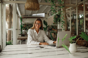 Smart woman in casual white clothes and eyeglasses sitting at wooden table in street cafe in front of laptop with coffee and documents and looking at camera