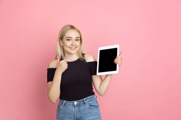 Naklejka premium Showing tablet, thumb up. Portrait of young woman with bright emotions on coral pink studio background. Blonde female model. Concept of human emotions, facial expression, sales, advertising, youth.
