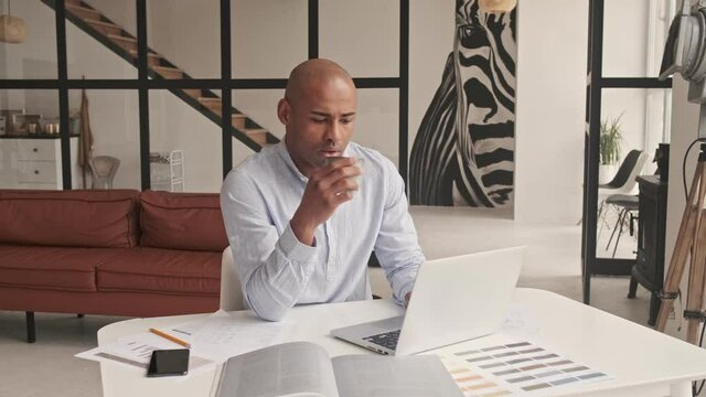 A handsome bald african american man is using his laptop while drinking coffee in the living room at home