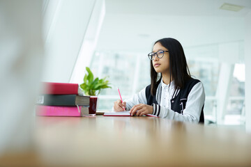 Pensive teen pupil in eyeglasses concentrate on creative home task writing ideas in notepad sitting...