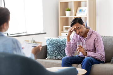 Obraz premium psychology, mental therapy and people concept - sad young indian man patient with glass of water and woman psychologist at psychotherapy session
