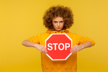 Portrait of angry curly-haired woman holding red Stop sign and looking at camera with negative...
