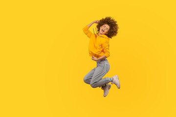 Portrait of overjoyed enthusiastic curly-haired girl in urban style hoodie and jeans jumping high...