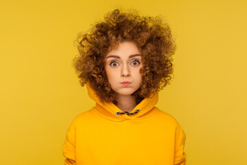 Portrait of cute curly-haired woman in urban style hoodie standing with puffed cheeks, holding...