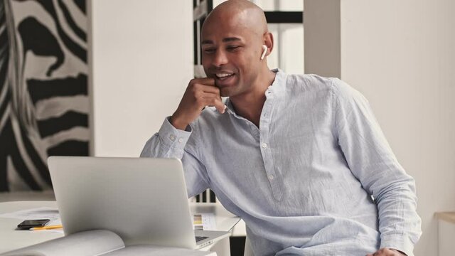 A cheerful good-looking african american man is talking with someone using video connection on his laptop in the living room at home