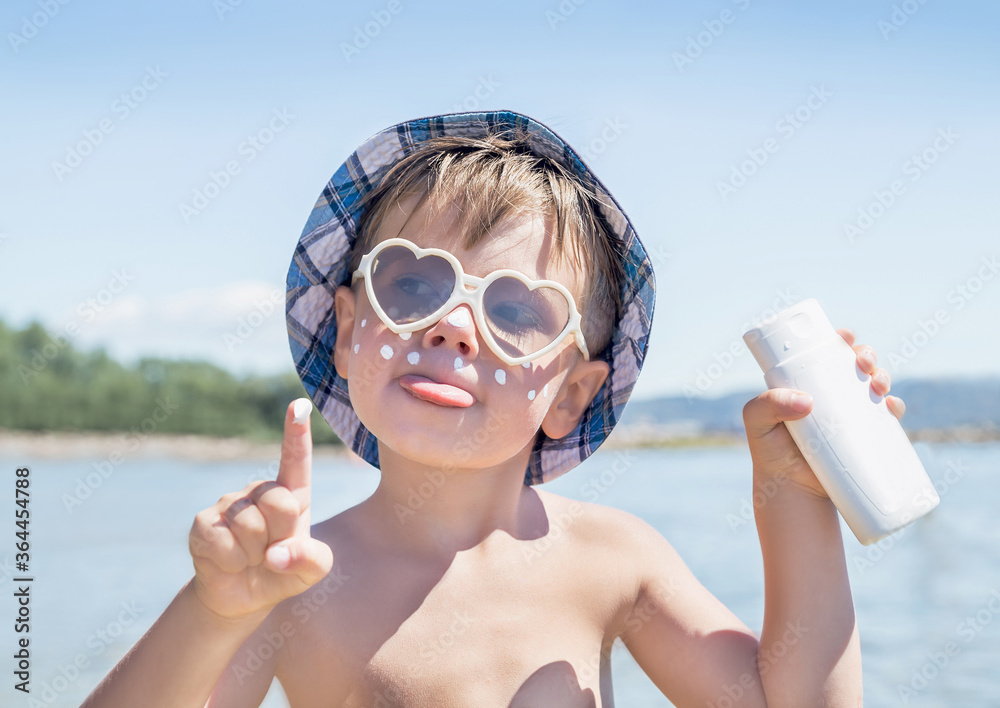 Wall mural Sunscreen is on hipster boy face before tanning during summer holiday on beach. Caucasian child (kid) have sunglasses, hat and holding container of suntan lotion (not far from Trieste, Italy).