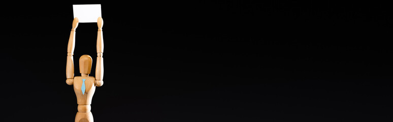 wooden marionette in tie holding blank placard isolated on black, panoramic shot