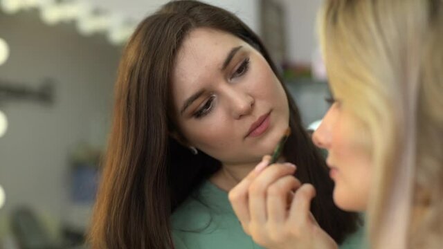 Side view of professional make-up artist applying eyeshadow to face of beautiful young woman in beauty salon, close-up. Concept of beauty, fashion and stylish makeup. Shooting in slow motion.