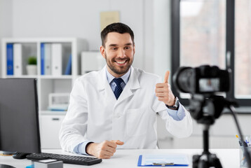 Naklejka na ściany i meble healthcare, medicine and blogging concept - happy smiling male doctor with camera recording video blog at hospital and showing thumbs up