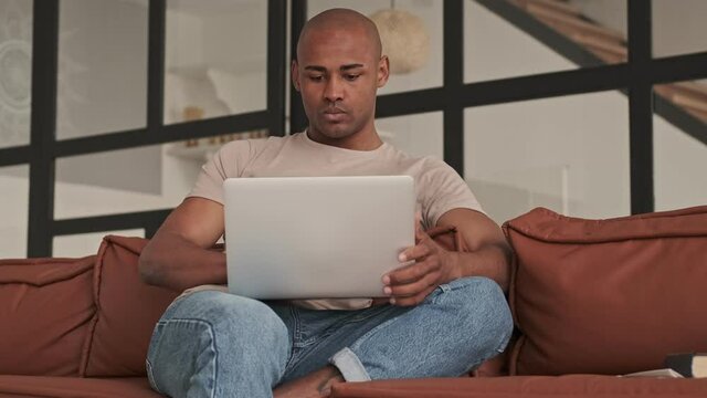 A serious african american man is using his laptop sitting on the couch in living room at home