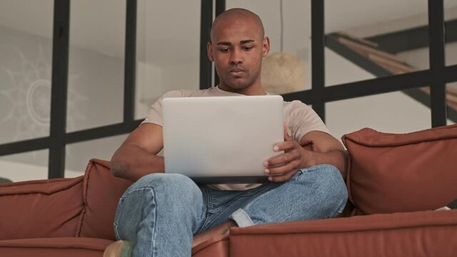 A concentrated african american man is using his laptop during working at home sitting on the couch in living room