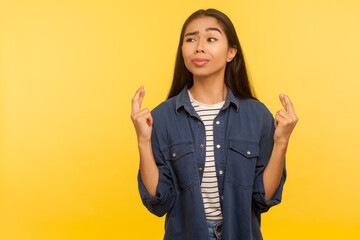 Portrait of girl making wish, holding fingers crossed for good luck, dreaming of lottery win, waiting success, in anticipation of miracle and fortune. indoor studio shot isolated on yellow background