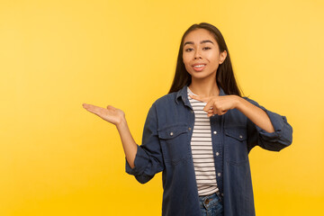 Attention, nice product advertise! Portrait of happy girl in denim shirt holding copy space with palm, pointing at empty place for commercial image. indoor studio shot isolated on yellow background