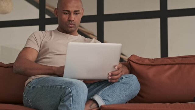 A calm african american man is using his laptop computer sitting on the sofa in living room at home