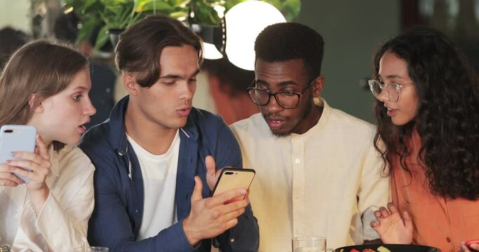 Crop view of millennial friends using phonest while browsing interne and sitting in cafe. Cheerful guy showing mobilephone content to coworkers and smiling. Concept of teamwork. Indoors.