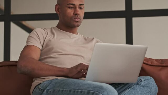 A handsome african american man is looking to his laptop sitting on the couch in living room at home