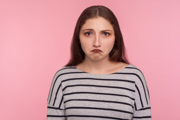 Fototapeta premium Sadness, negative emotions. Portrait of unhappy depressed young woman in striped sweatshirt looking with disappointed expression, resentful grimace. indoor studio shot isolated on pink background