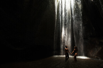 Beautiful couple in a cave with a waterfall. Athletic man and woman under the streams of a waterfall. A sexy couple under a tropical waterfall in twilight light. Tukad Chepung Waterfall. Copy space