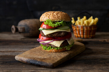 Hamburger with tomatoes, onions, cucumber, lettuce and melting cheese served on a  rustic wooden board with french fries and vegetables in background
