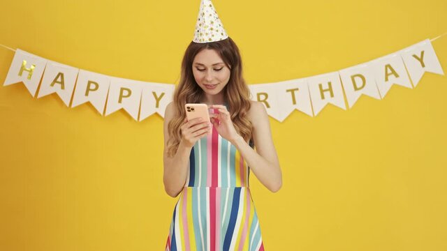 A lovely woman is watching something cute on her smartphone on a birthday party isolated over a yellow background