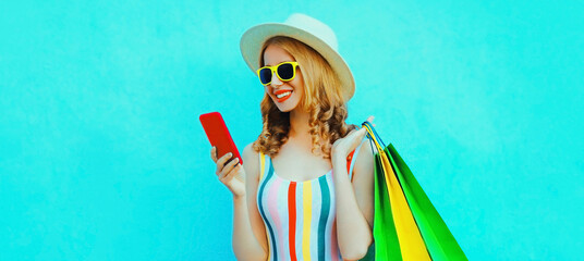 Portrait of smiling young woman with phone and shopping bags in colorful striped t-shirt, summer round hat on blue background