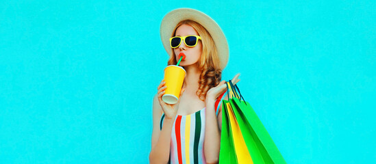 Portrait of young woman drinking juice with shopping bags in colorful striped t-shirt, summer round hat on blue background