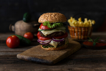 Hamburger with tomatoes, onions, cucumber, lettuce and melting cheese served on a  rustic wooden board with french fries and vegetables in background