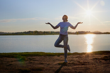 Fototapeta na wymiar Silhouette of a girl doing sports exercises on the lake and sunset with the sun and rays in the background. Female yogi meditates during yoga.