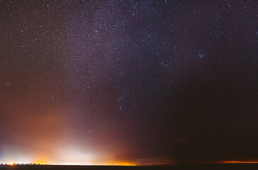 Night Starry Sky With Glowing Stars Above Dark Dround. Bright Glow Of Stars. Illumination Lights Of Small Town Or Village. Milky Way Galaxy