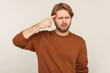 Idiotic plan! Portrait of bearded man showing stupid gesture, pointing finger to temple and looking dissatisfied with reckless silly idea, dumb plan. indoor studio shot isolated on gray background