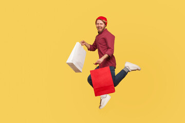Lively happy hipster trendy guy running in air with shopping bags in hands, hurrying to catch sale, flying and rushing for thrift discount. full length studio shot isolated on yellow background
