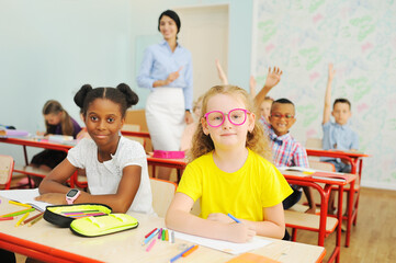 two children-an African-American girl and a Caucasian girl in pink glasses are smiling sitting at a Desk in a primary school against the background of a teacher 