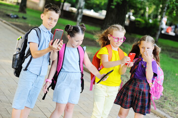Obraz premium a group of school children in colorful clothes with school bags with smartphones in their hands go to school. Back-to-school gadgets, the day of knowledge, dependence on the mobile phone.