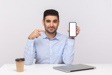 Cheerful elegant young businessman sitting in office workplace, pointing at mobile phone with empty screen, mock up blank display for advertisement. indoor studio shot isolated on white background