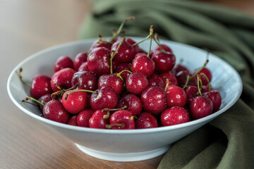 Red cherries in a white bowl on a wooden table with a green shawl , close up, still life photography