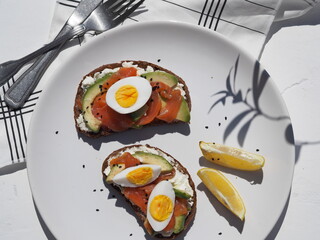 Sandwiches with whole grain bread, cheese, fish and an egg in a white plate on a napkin on a white wooden background.The shadow of the plant.