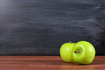 Two fresh green apples on brown wooden desk with blackboard texture in background. Copy space, close-up. School snacks or organic food concept