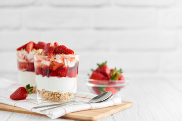 Homemade layered dessert with fresh strawberries, cream cheese or yogurt, granola and strawberry jam in glasses on white wood background. Healthy organic breakfast or snack concept. Selective focus.