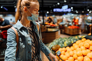young woman in a medical protective mask on her face, buying fruit in a supermarket. a young lady buys oranges at a grocery store. Sale, shopping, food, coronavirus