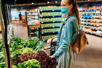 young woman in a medical protective mask on her face buys vegetables and fruits in a supermarket. a young lady buys vegetables and herbs at a grocery store. Sales, purchases, food, coronavirus