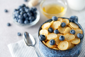 Blue Bowl of mini pancake cereal with blueberries, maple syrup and milk on concrete background. Tiny pancakes, new food trend concept. Cute healthy breakfast or snack. Recipe, menu.