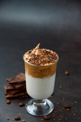 Glass of trendy fluffy creamy dalgona or whipped instant coffee with milk, coffee beans and chocolate on the dark background. New popular food and drink trend concept. Copy space.