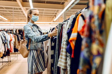 Obraz na płótnie Canvas girl in a medical protective mask on her face, chooses new clothes in the store and smiles. Shopping, quarantine, coronavirus