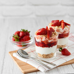 Homemade layered dessert with fresh strawberries, cream cheese or yogurt, granola and strawberry jam in glasses on white wood background. Healthy organic breakfast or snack concept. Selective focus.