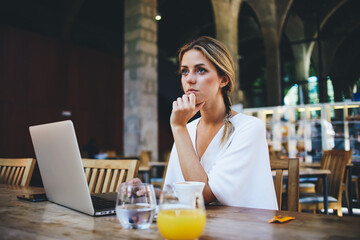 Portrait of a young women thinking about something while sitting front portable laptop computer in modern cafe interior, beautiful dreamy female taking break between work on net-book during breakfast