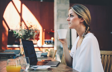 Beautiful dreamy female holding cup of coffee while sitting in front of open portable laptop computer, young thoughtful businesswomen enjoying hot beverage while relaxing after work on her net-book